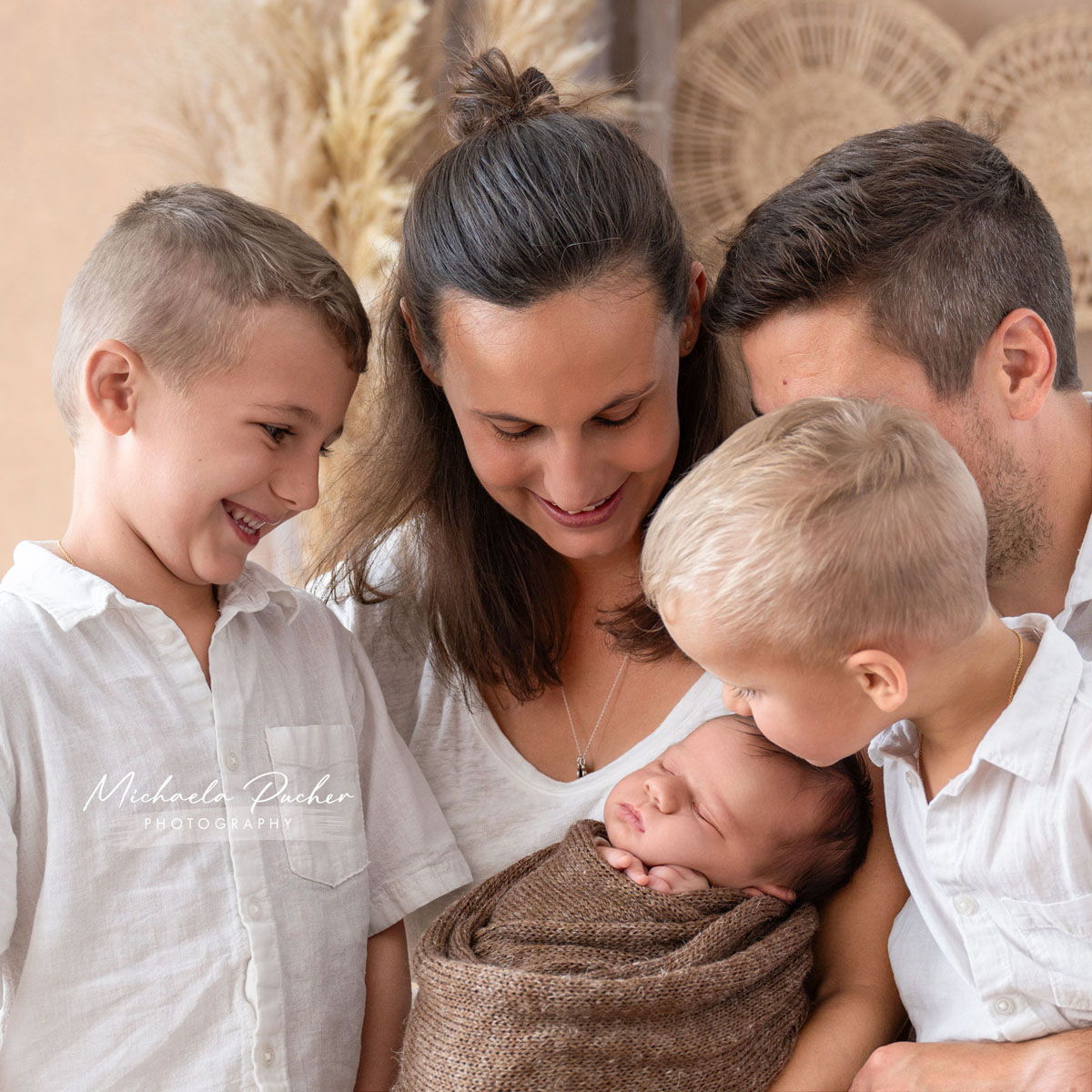 Familie mit drei Kindern: Die Eltern halten das Neugeborene, während die beiden älteren Brüder liebevoll und neugierig auf das Baby blicken.